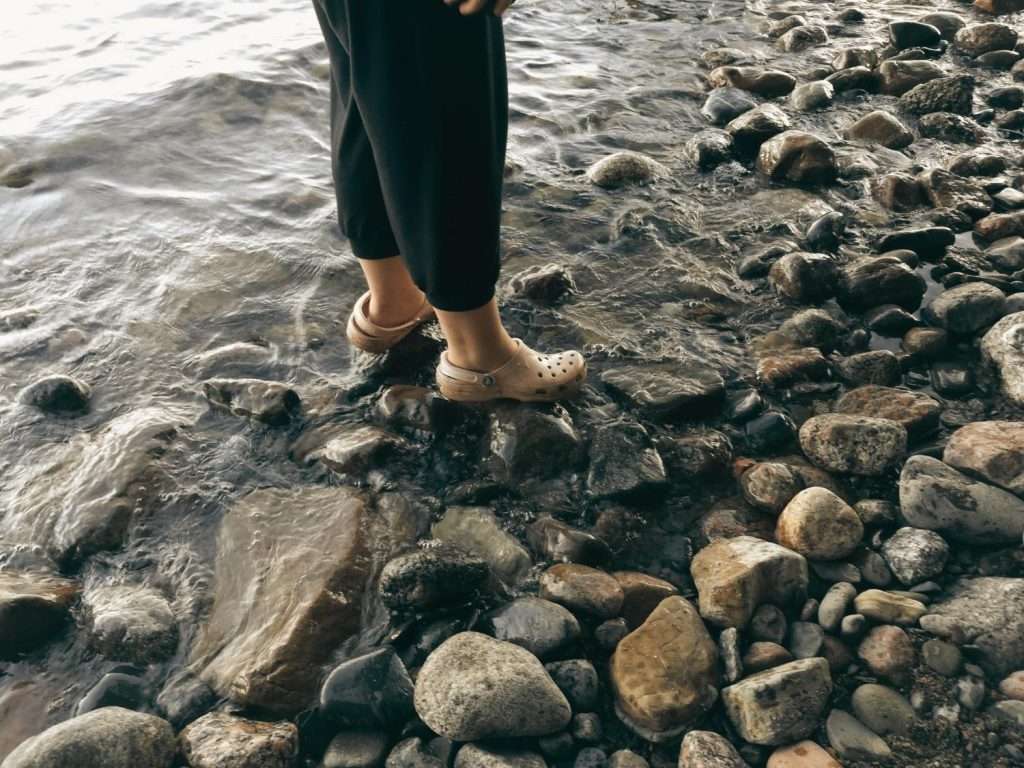 Mensch steht am Rand eines Gewässers in Sandalen im Wasser